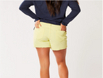 Back view of female model wearing Carve Designs Oahu shorts in citron (yellow). Shorts are corduroy with 4" inseam, front button/zipper closure, two back pockets, and raw hem bottom. Paired with navy blue long sleeve top.